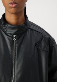 Veste en cuir noir avec un col haut et une fermeture éclair. Détails texturés remarquables sur les épaules et un logo brodé sur la poitrine.