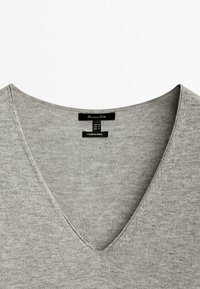 Pull en maille grise avec un design en V. Il se distingue par sa texture douce et un label en détail au niveau du col, fabriqué à partir d'un tissu doux.