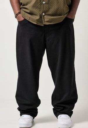 Person trägt lockere schwarze Hose, weiße Turnschuhe, grünes Hemd zum Knöpfen und ein Perlenarmband, die Hände in den Taschen.