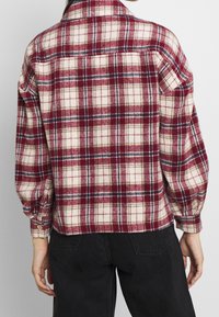 Koszula flanelowa w kratę w kolorach kremowym, czerwonym i granatowym, z mankietem na guziki, prostym krojem i widocznymi szwami, o miękkiej fakturze.