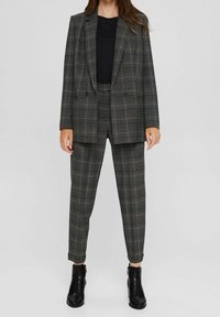 Costume à carreaux en tissu tissé, présentant une palette de couleurs grises et brunes. La veste est croisée, à coupe ample, associée à un pantalon fuselé.