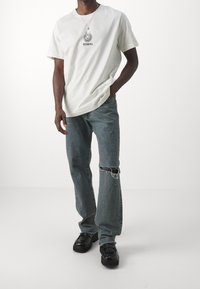 Biała bawełniana koszulka z graficznym logo na środku, w zestawieniu z jasnoniebieskimi podartymi dżinsami i czarnymi skórzanymi butami. Prosty codzienny strój.