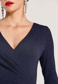 Frau in einem navyblauen glitzernden Kleid mit V-Ausschnitt, kräftigem roten Lippenstift und hängenden runden Kristallohrringen, vor einem neutralen Hintergrund.