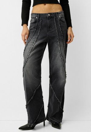Personne portant un jean noir taille haute, jambes larges avec des coutures diagonales texturées et des escarpins noirs à bout pointu, montrée de la taille aux pieds.