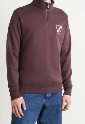 Homme portant un pull à fermeture éclair quart de zip bordeaux avec un logo blanc en forme de bouclier sur la poitrine gauche et un jean bleu, debout devant un fond uni.
