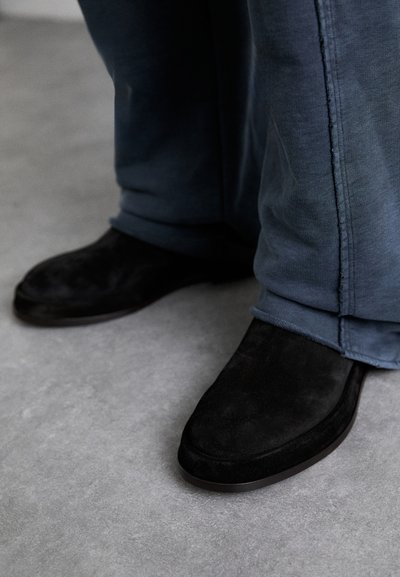 Pantofi negri din suede, cu vârful rotunjit și talpă plată, purtați cu pantaloni din denim albastru, cu croială largă, având un tiv brut.  Materialul texturat este vizibil.