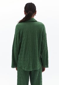 Outfit verde texture che presenta una camicia morbida con colletto e maniche lunghe, abbinata a pantaloni coordinati, il tutto realizzato in materiale leggero.