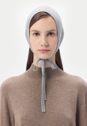 Junge Frau mit grauer Strickmütze, die unter dem Kinn gebunden ist, und einem braunen Rollkragenpullover, die nach vorne blickt mit neutralem Gesichtsausdruck.