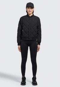 Zwart gewatteerd bomberjack met een ronde kraag, drukknoopsluiting aan de voorkant en ribgebreide mouwen, gecombineerd met zwarte leggings en sneakers.