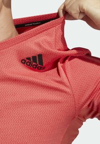 Rød atletisk skjorte med et struktureret mønster, der har et sort Adidas-logo på brystet og en tætsiddende design.