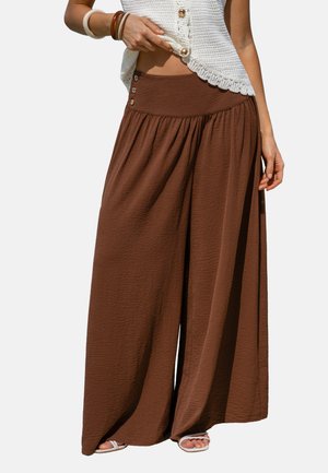 Vrouw draagt wijde bruine broek met hoge taille en knopen, gecombineerd met witte sandalen en een mouwloze gebreide top.