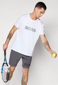 Λευκό αθλητικό μπλουζάκι με το "BORG" σε γκρι χρώμα στο στήθος, συνδυασμένο με γκρι σορτς που έχουν το μαύρο λογότυπο "BORG". Ο παίκτης κρατά μια μπάλα τένις.