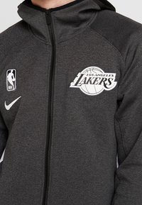 Grå Nike zip-up jakke med Los Angeles Lakers logo, tekstureret stof, høj krave og sorte accenter.