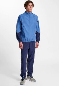 Sportstræningsdragt bestående af en lyseblå jakke med sorte detaljer og matchende mørkeblå bukser, med en slank pasform og elastiske manchetter.