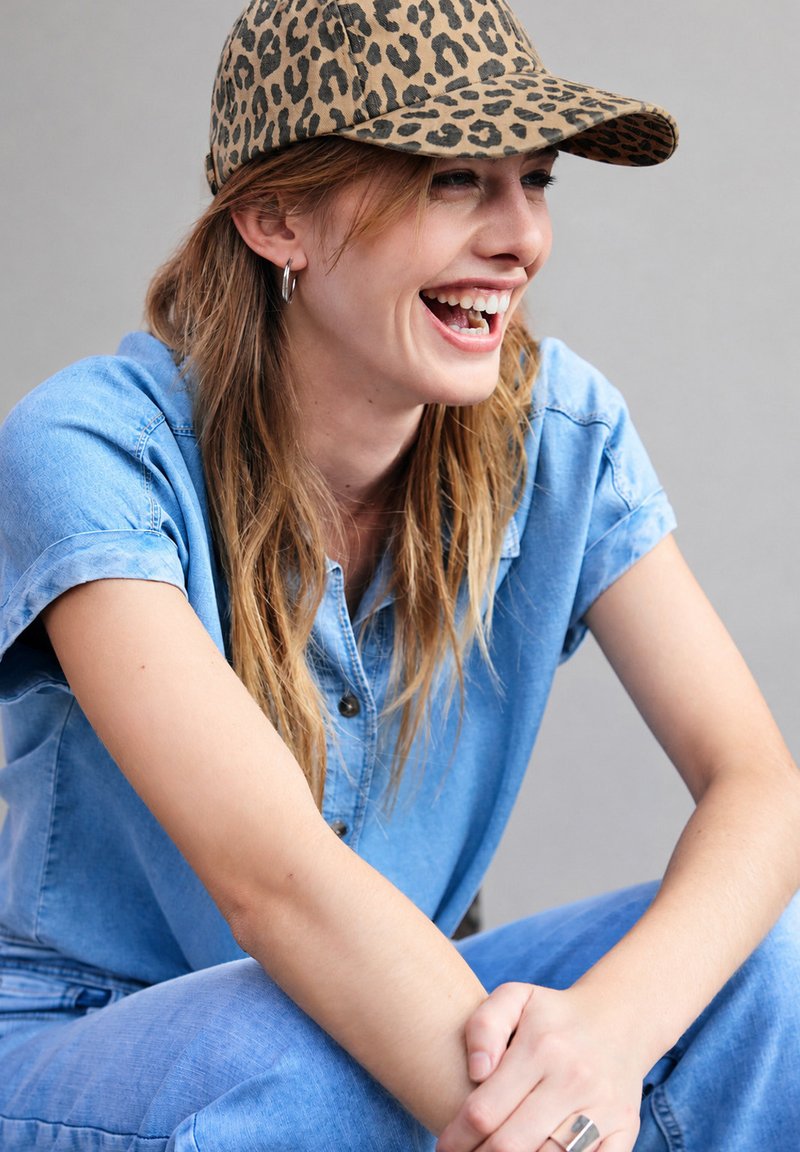 Jeune femme en tenue en denim bleu et casquette à motif léopard souriante, assise les bras croisés sur les genoux devant un fond uni.