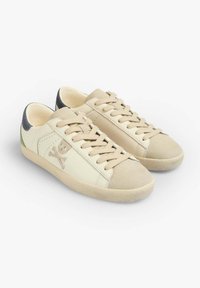 Baskets beige avec une tige en cuir lisse, un capuchon en suede et des accents verts et bleus marine. Présente un logo de crâne et os croisés sur le côté.
