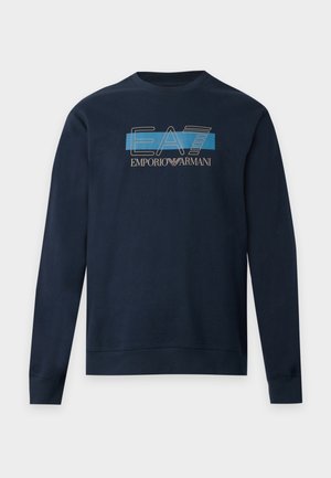 Un mannequin aux cheveux courts décolorés porte un sweat-shirt bleu marine Emporio Armani EA7 et un pantalon assorti sur un fond clair uni.