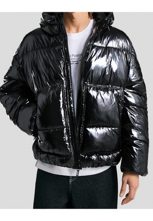 Mann trägt glänzende schwarze Steppjacke mit Kapuze über weißem, bedrucktem T-Shirt und dunklen Jeans, Hände in den Taschen.
