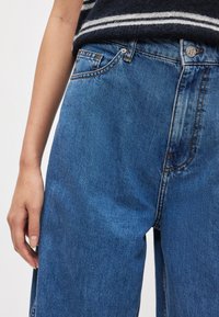 Μπλε τζιν παντελόνια με φαρδιά μπατζάκια διαθέτουν μέση μεσαίου ύψους, κλασικό σχέδιο με πέντε τσέπες και αντίθετη ραφή. Το ύφασμα έχει ομαλή υφή.