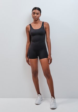 Une femme est debout contre un mur uni, portant une combinaison noire sans manches ajustée et des baskets blanches, regardant devant elle avec une expression neutre.