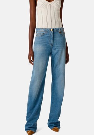 Donna che indossa jeans azzurri a gamba dritta, top bianco lavorato a maglia senza maniche e scarpe marroni con punta su sfondo semplice.