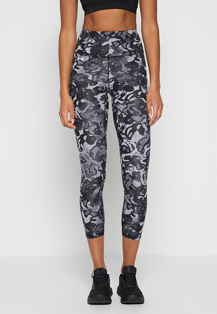 Femme portant un legging cropped noir à motif camouflage, un soutien-gorge de sport noir et des baskets noires, se tenant devant un fond clair uni.