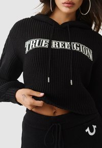 Schwarzer, gerippter Hoodie mit "TRUE RELIGION"-Logo in Weiß, ausgestattet mit Kapuze und Kordelzug sowie einem verkürzten Schnitt. Kombiniert mit schwarzen, gerippten Hosen.