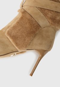 Botine din suede bej și blană artificială, cu vârf ascuțit și toc subțire de tip stiletto, pe fundal alb.