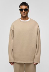 Beige oversized sweatshirt van zachte stof met een ronde hals, lange mouwen en een afgewerkte zoom. Draagt zwart smal zonnebril.
