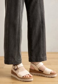 Beige wedge sandals with wide straps and a round buckle detail. Cork-textured midsole and textured white rubber sole. Gray wide-leg pants.