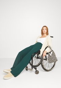 Femme aux cheveux châtain clair en fauteuil roulant portant un haut blanc, un cardigan crème, un pantalon vert, des chaussures beiges, tenant un sac à carreaux noir et blanc.