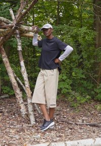 Svart kortermet skjorte, beige knelange shorts med "TERREX"-logo, og grå sneakers. Står i et skogområde med trær.