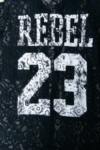 Tessuto in pizzo nero con motivo floreale, con testo in grassetto bianco che recita "REBEL 23" in uno stile vissuto e irregolare.
