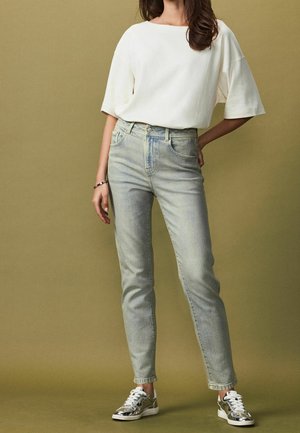 Jean en denim clair avec une coupe fuselée, associé à un haut blanc à manches courtes ample et des baskets argentées sur un fond olive atténué.