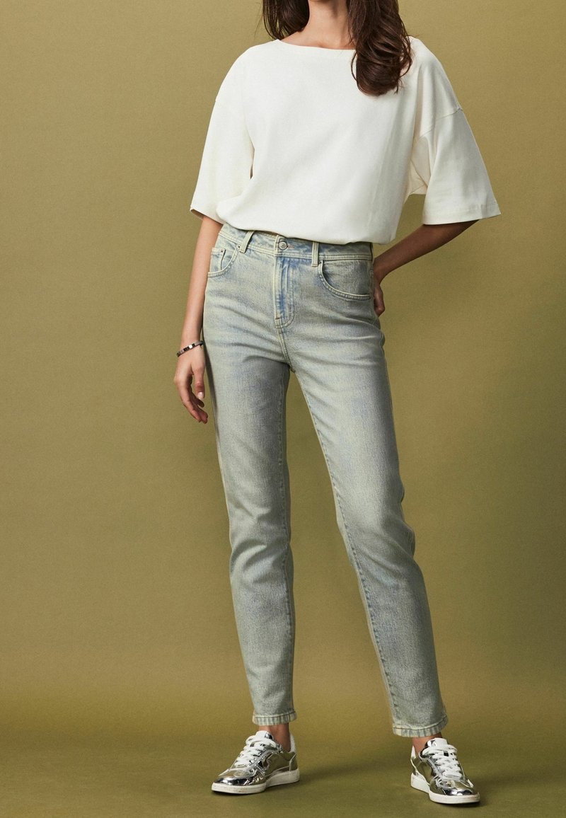 Jeans in denim chiaro con vestibilità affusolata, abbinati a una maglietta bianca a maniche corte e scarpe da ginnastica argentate su uno sfondo verde oliva tenue.