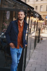 Laivansininen vedenpitävä takki nappitaskuilla, oranssin paidan päällä, vaaleansinisten farkkujen kanssa, seisoo lasiseinää vasten.