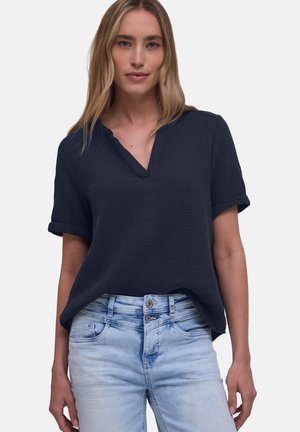 Femme portant un chemisier ample à manches courtes bleu marine foncé et un jean bleu clair à double bouton devant un fond uni.