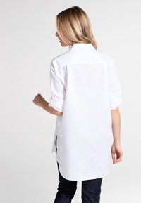 Camicia bianca a maniche lunghe in tessuto leggero, con maniche arrotolate e orlo arrotondato, indossata con jeans scuri.