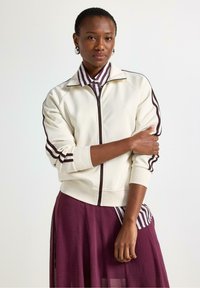 Krémová zipová bunda s vínovými pruhy na rukávech. Nositá přes pruhovanou košili s límečkem, zkombinovaná s volnou vínovou sukní.