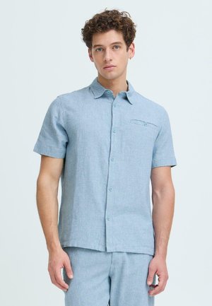 Jeune homme aux cheveux bouclés portant une chemise à manches courtes bleu clair boutonnée et un pantalon assorti, debout devant un fond uni.