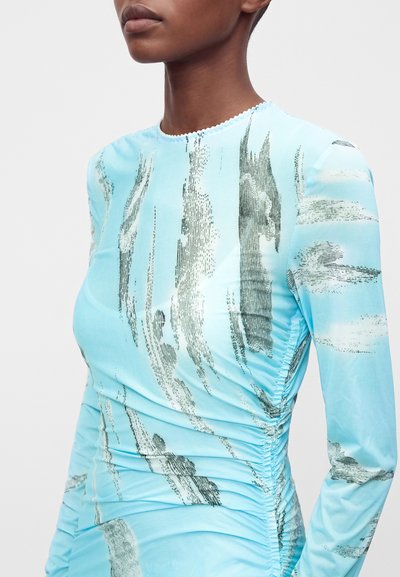 Personne portant un haut à manches longues bleu clair avec des motifs abstraits de coups de pinceau verticaux noirs et blancs et des côtés froncés.