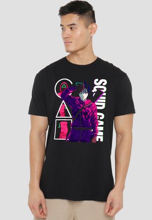 Young man in a black T-shirt featuring a colorful "Squid Game" graphic, striking a pose with a hand on his forehead. Neutral background.