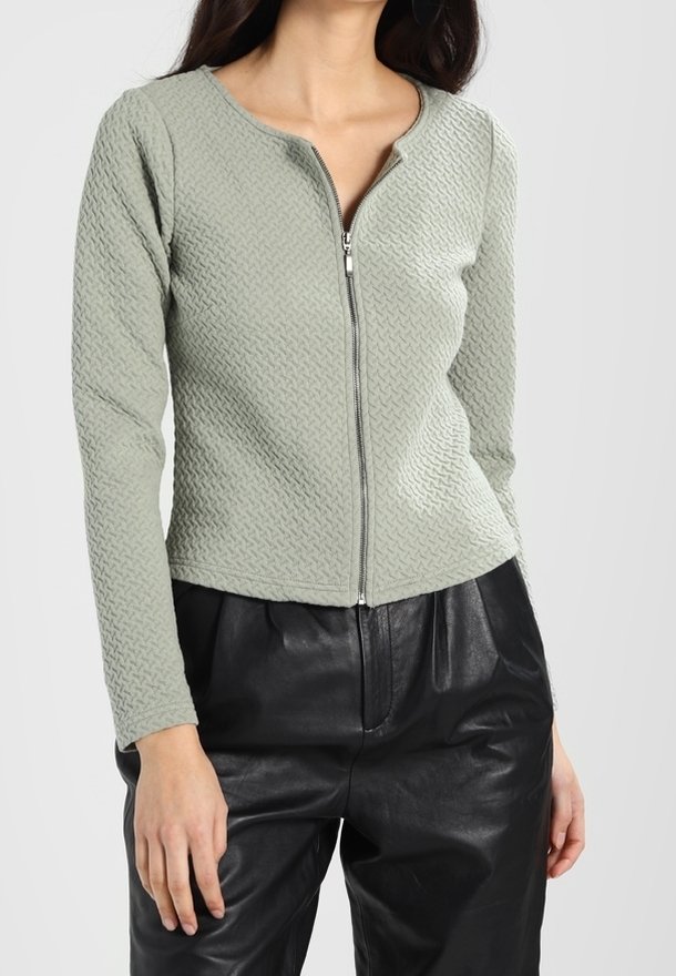 Femme portant un top zippé texturé vert clair à manches longues et un pantalon en cuir noir taille haute sur un fond uni.