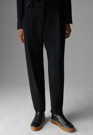 Persoon draagt zwarte taps toelopende broek en zwarte leren platformsneakers met dikke bruine zolen, staand op een grijze vloer.