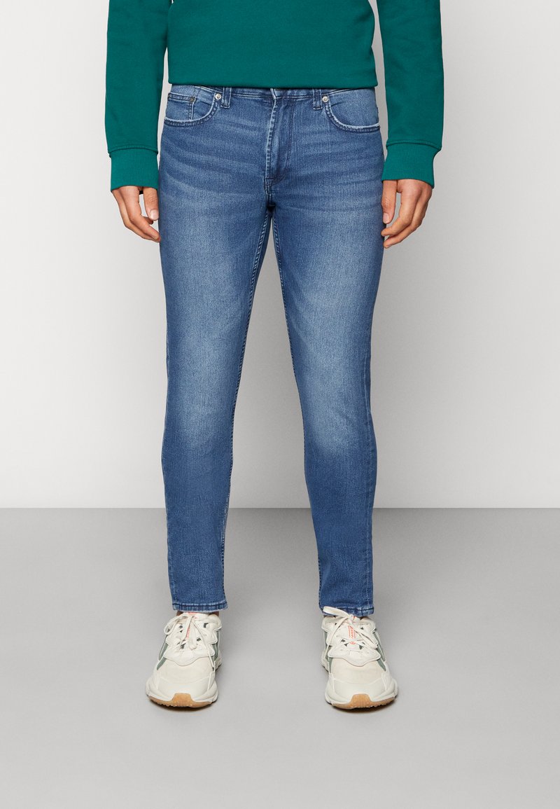 Blå smale jeans med en falmet vask, frontlommer og en standard livlinje, sammen med hvite og grå sneakers.