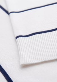 Tessuto bianco con strisce orizzontali blu navy; polsino a coste visibile. La texture liscia evidenzia il design pulito e il contrasto di colori.