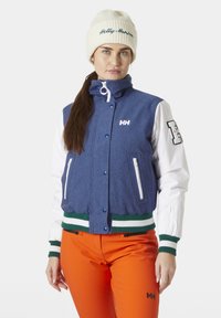 Damesjas met een blauwe romp, witte mouwen, groene en witte gestreepte afwerking, met ritszakken en geborduurd logo. Gecombineerd met oranje broeken.