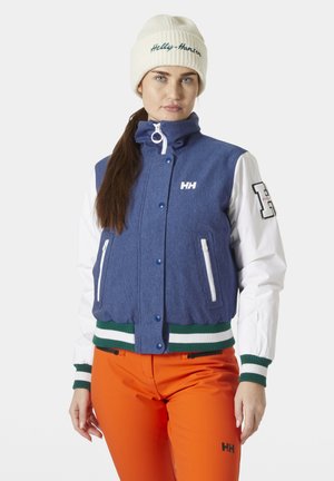 Damesjas met een blauwe romp, witte mouwen, groene en witte gestreepte afwerking, met ritszakken en geborduurd logo. Gecombineerd met oranje broeken.