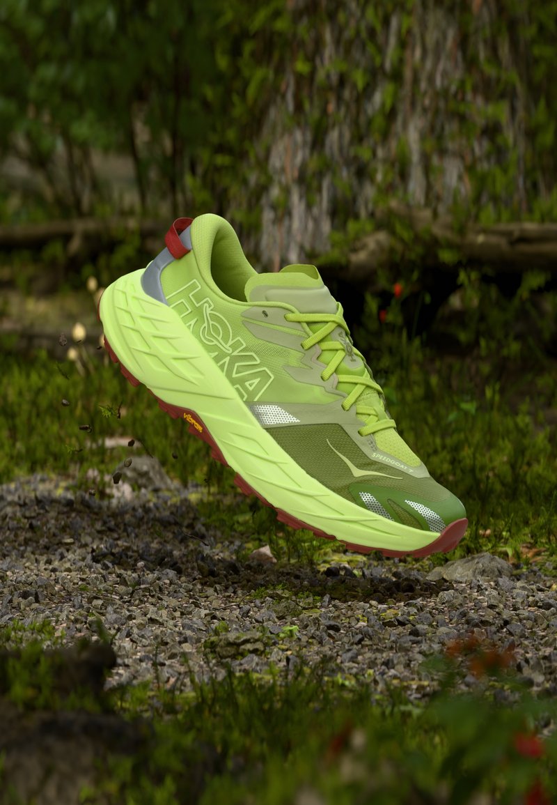 Πράσινο παπούτσι τρεξίματος με υφασμάτινο επάνω μέρος με υφή, αντανακλαστικές λεπτομέρειες και χοντρή σόλα. Διαθέτει κόκκινες και γκρι λεπτομέρειες, σχεδιασμένο για εξωτερική χρήση.