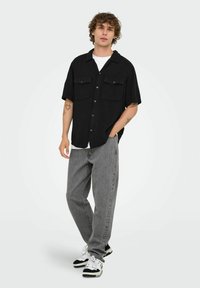 Sort kortærmet skjorte med to brystlommer, parret med lysegrå baggy jeans og sort-hvide sneakers, sat på en ensfarvet baggrund.